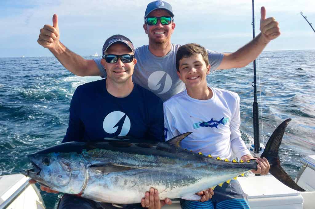 Three smiling anglers sitting on a boat, holding a large Tuna on a sunny day