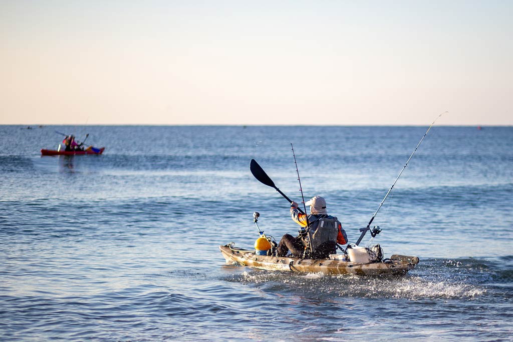 A kayak fisherman rowing in open waters while another kayak with multiple guests is visible in the distance