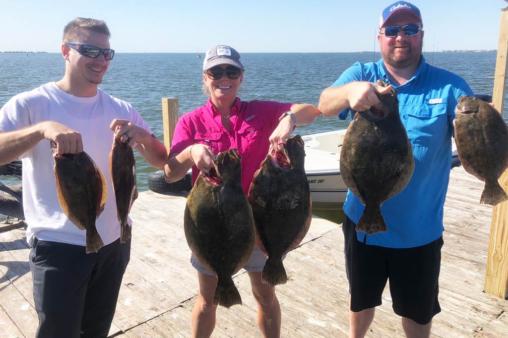 Two men and a woman standing on a dock holding two Flounder each on a sunny day