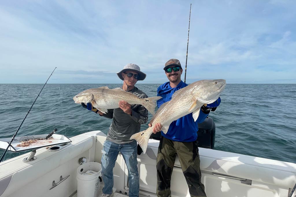 Two fishermen standing on a charter boat, holding two big Redfish with blue skies and water in the background
