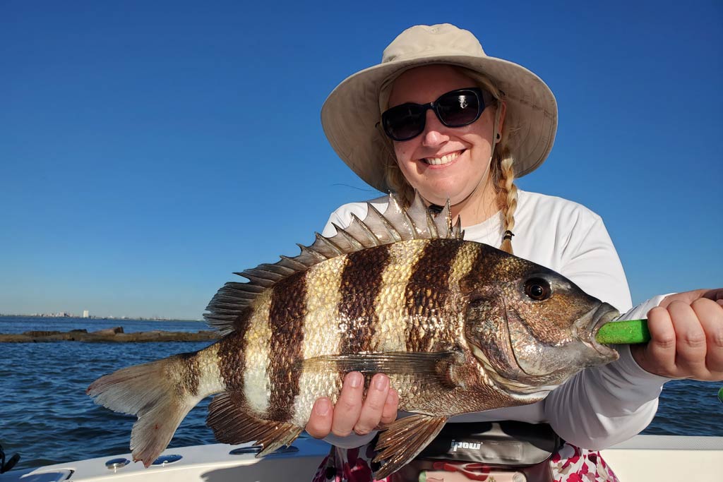 A smiling woman in a hat and sunglasses holding a Sheepshead, with blue skies in the background