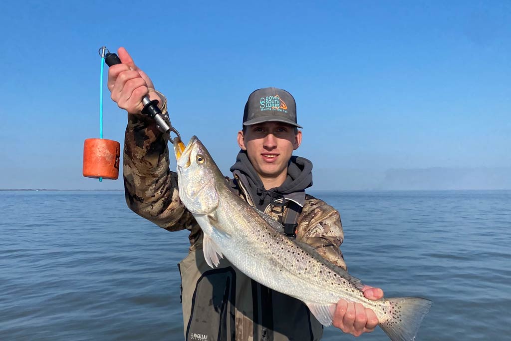 A young angler in a cap holding a big Speckled Trout with blue skies and water behind him