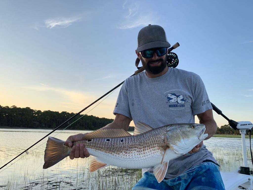 A photo of a fly fisherman with a fly fishing rod behind him and a Redfish in front of him caught while fly fishing in Jekyll Island’s inshore waters