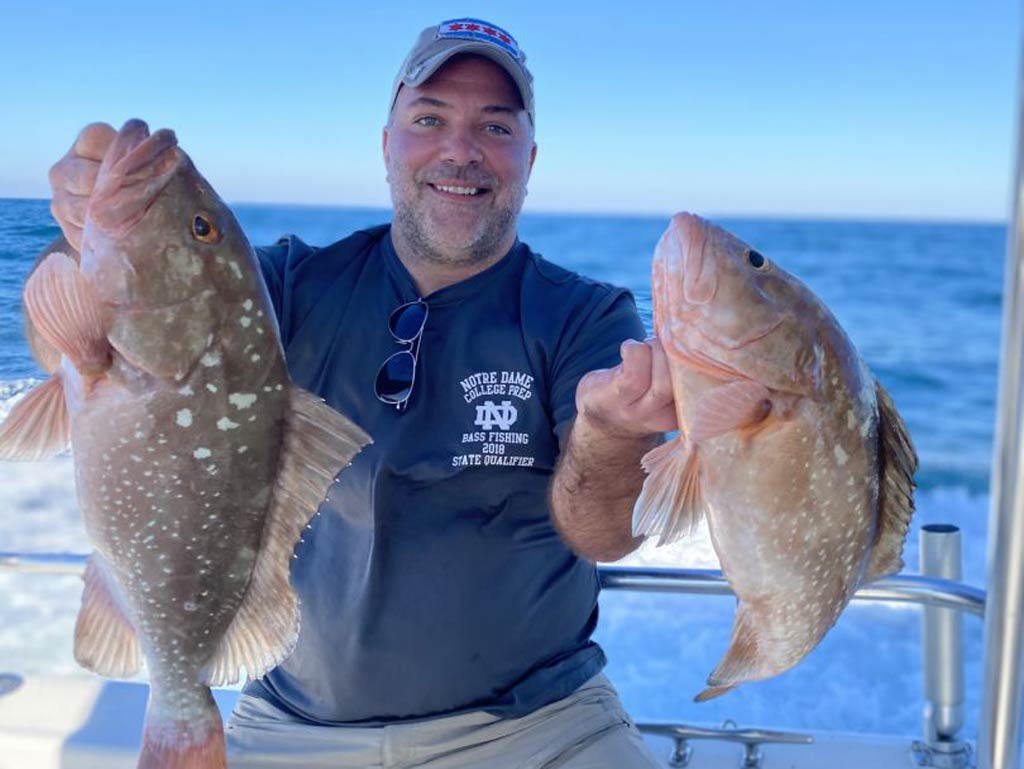 A happy angler holding two Groupers, one in each hand, while standing on a Madeira Beach fishing charter