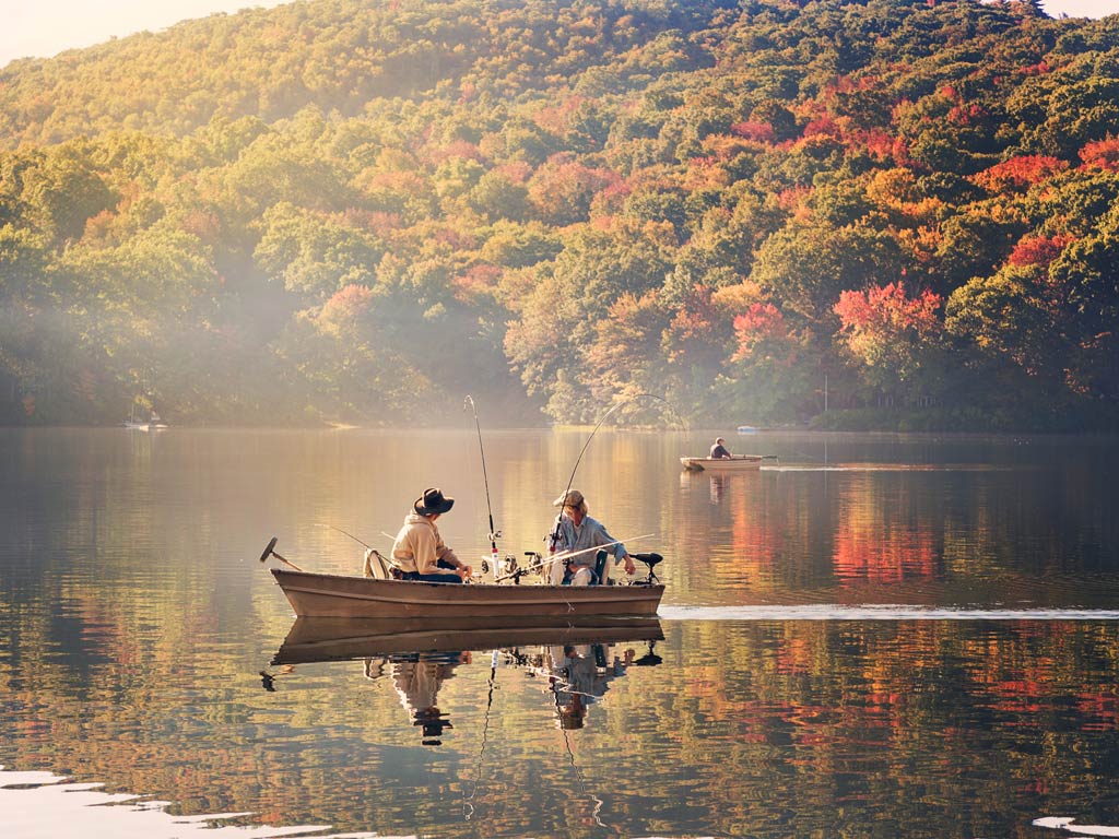 A photo of three anglers fishing from two boats on a lake during a sunny fall day, with green and amber foliage in the distance