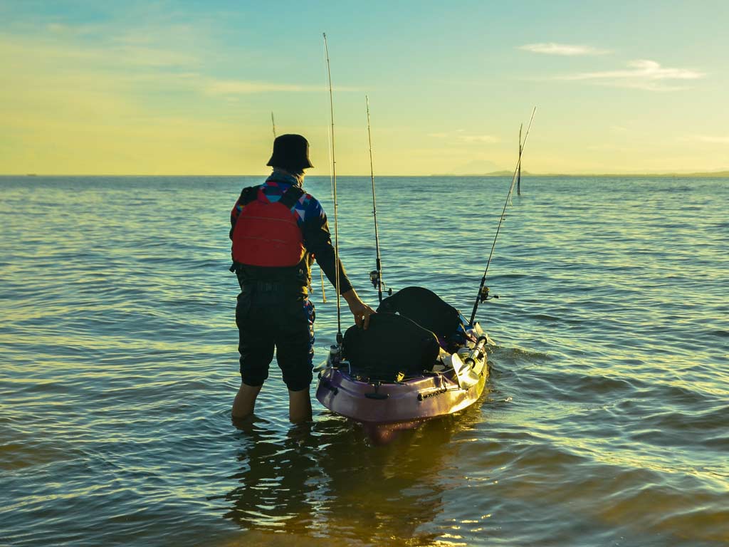 An fisherman standing in shallow waters with his kayak next to him.