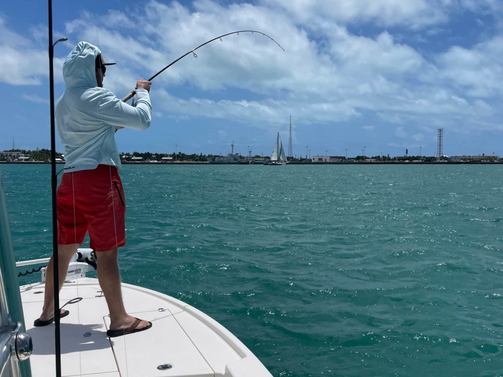 A picture showing a male angler wearing red shorts, flip-flops, and a light blue hoodie, holding a fishing rod and standing on a fishing boat with Key West in the background