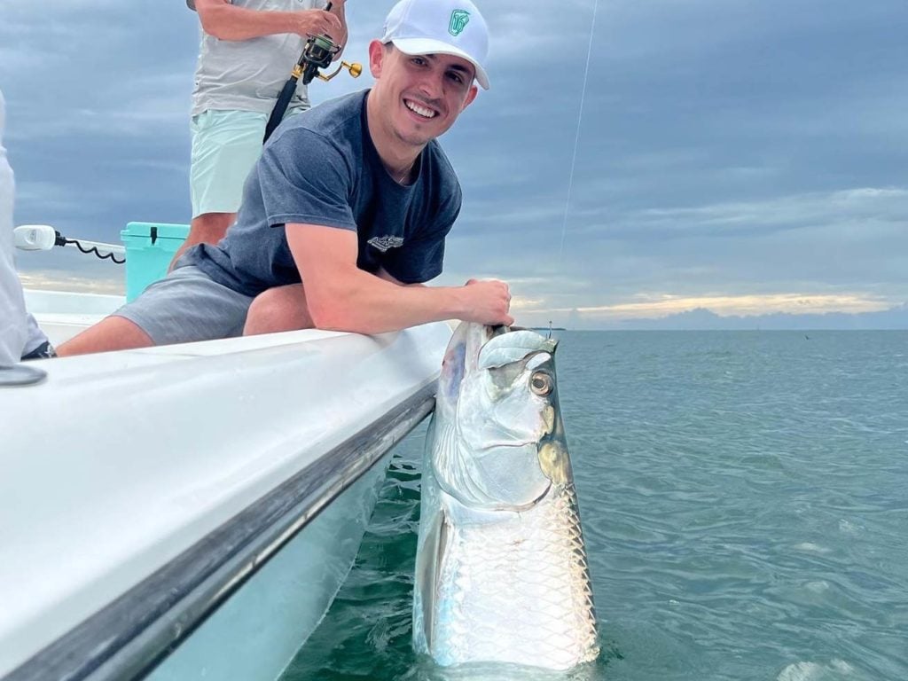 Two anglers on a fishing boat in Key West, Florida, one of them holding a fishing rod, while the other iss smiling and holding a large Tarpon in the water