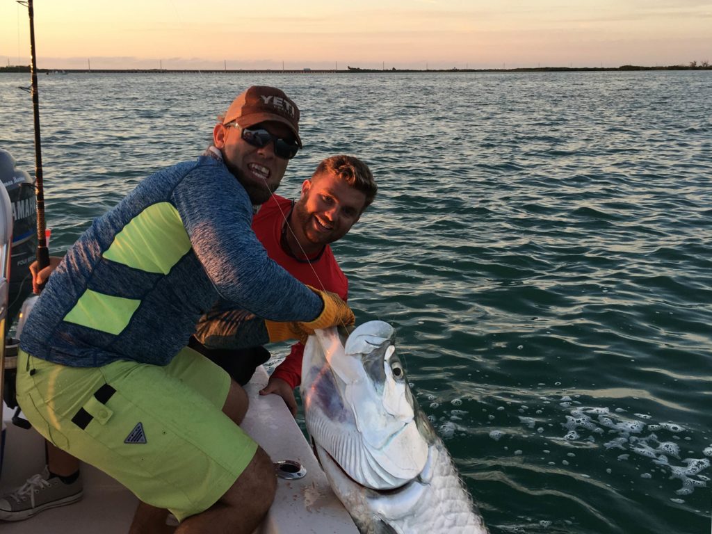 Two male anglers, one in a red t-shirt holding a fishing rod, and the other wearing a Yeti cap and dark sunglasses, holding a large Tarpon in the water from a boat by the shore in Key West, Florida