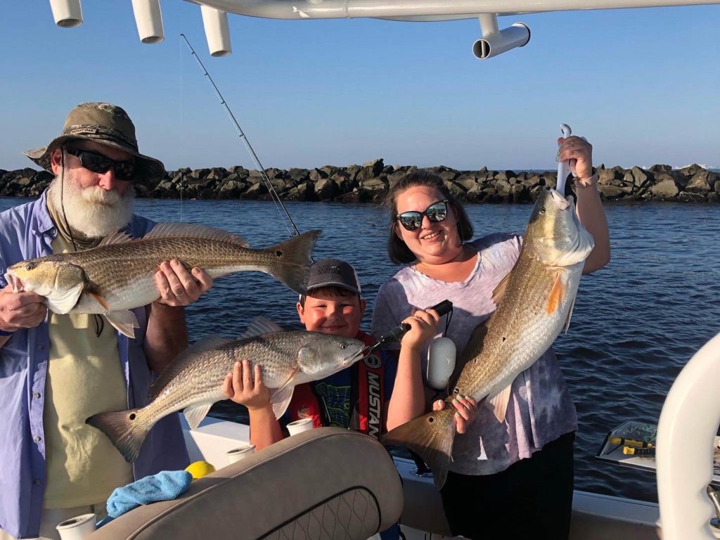 Three anglers of different ages – one female and two males – each holding a freshly caught Redfish, standing on a fishing boat in North Myrtle Beach, South Carolina.