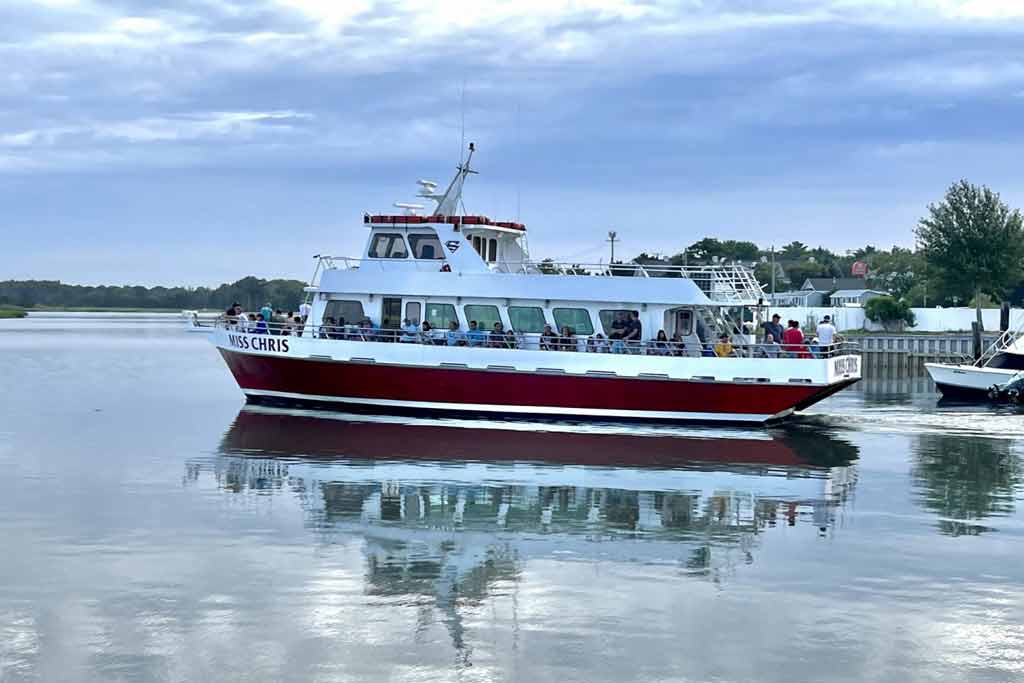 A dark red and white party fishing boat on the water with anglers on it on a cloudy day