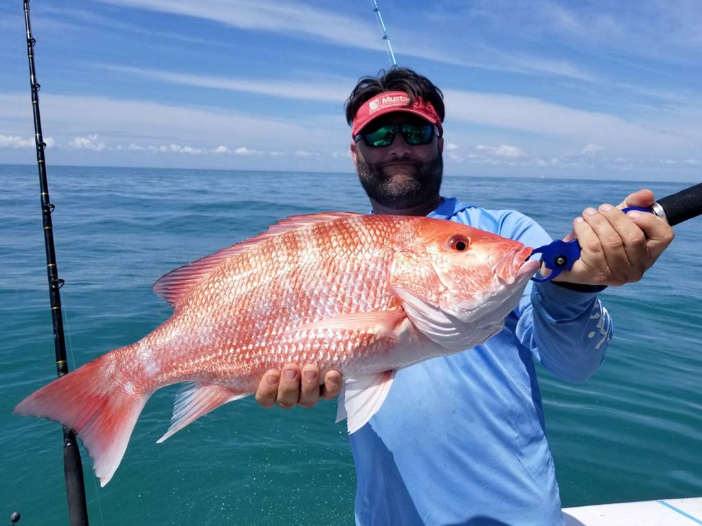 An angler posing with a big Red Snapper he reeled in, with calm ocean waters behind him.