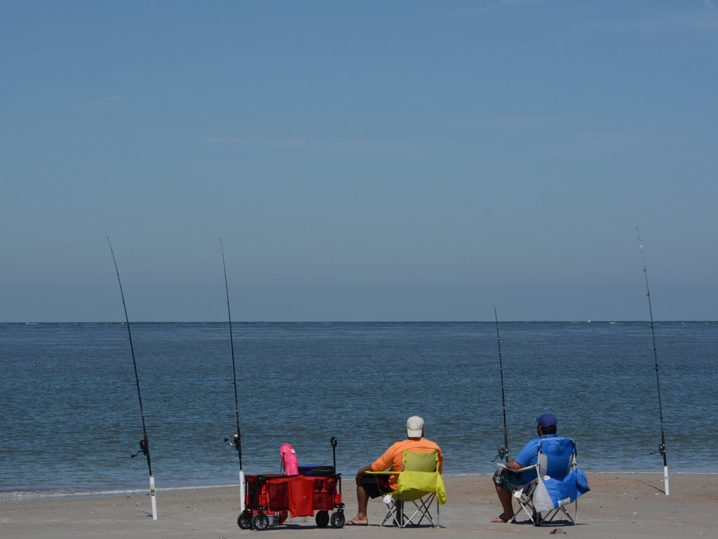 Two anglers sitting in chairs on a beach in Fernandina Beach, with several fishing rods beside them.