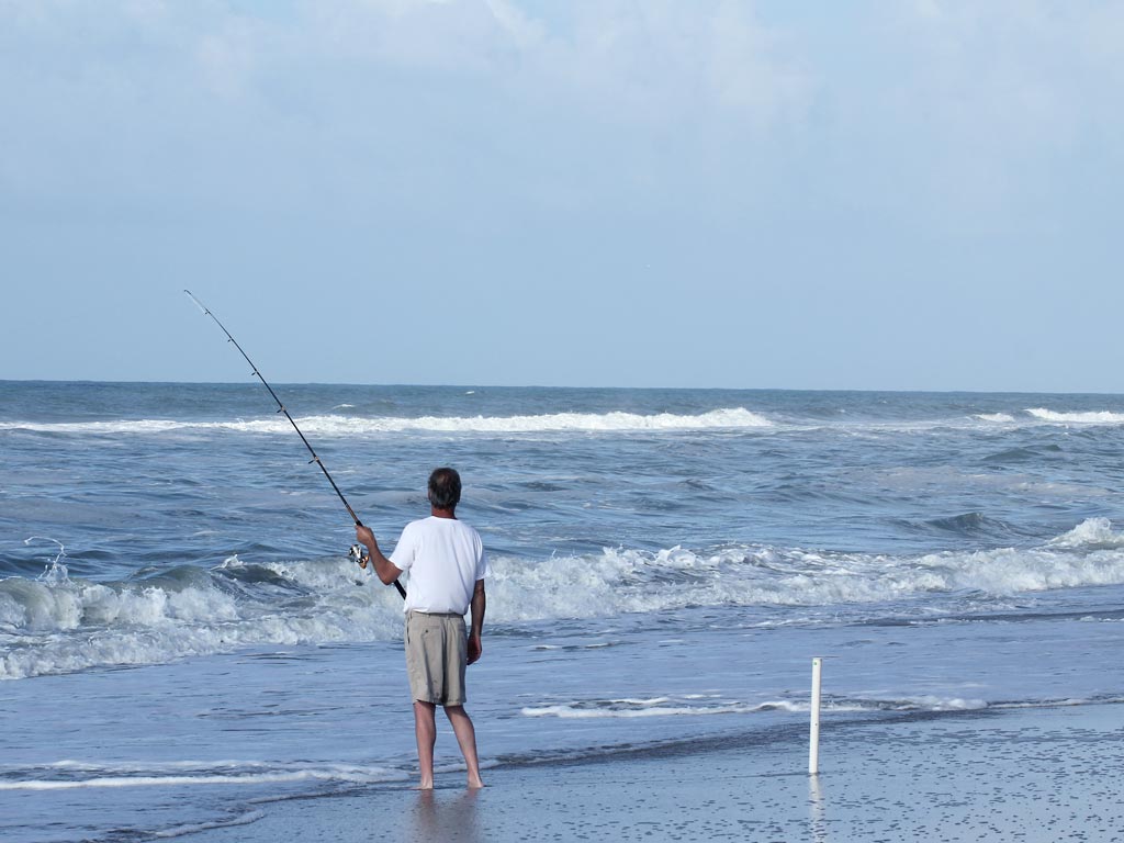 An angler on a beach, turned towards the ocean and holding a fishing rod.