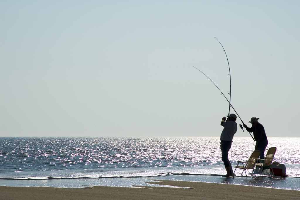 Two fishermen casting their fishing lines from a beach into the surf