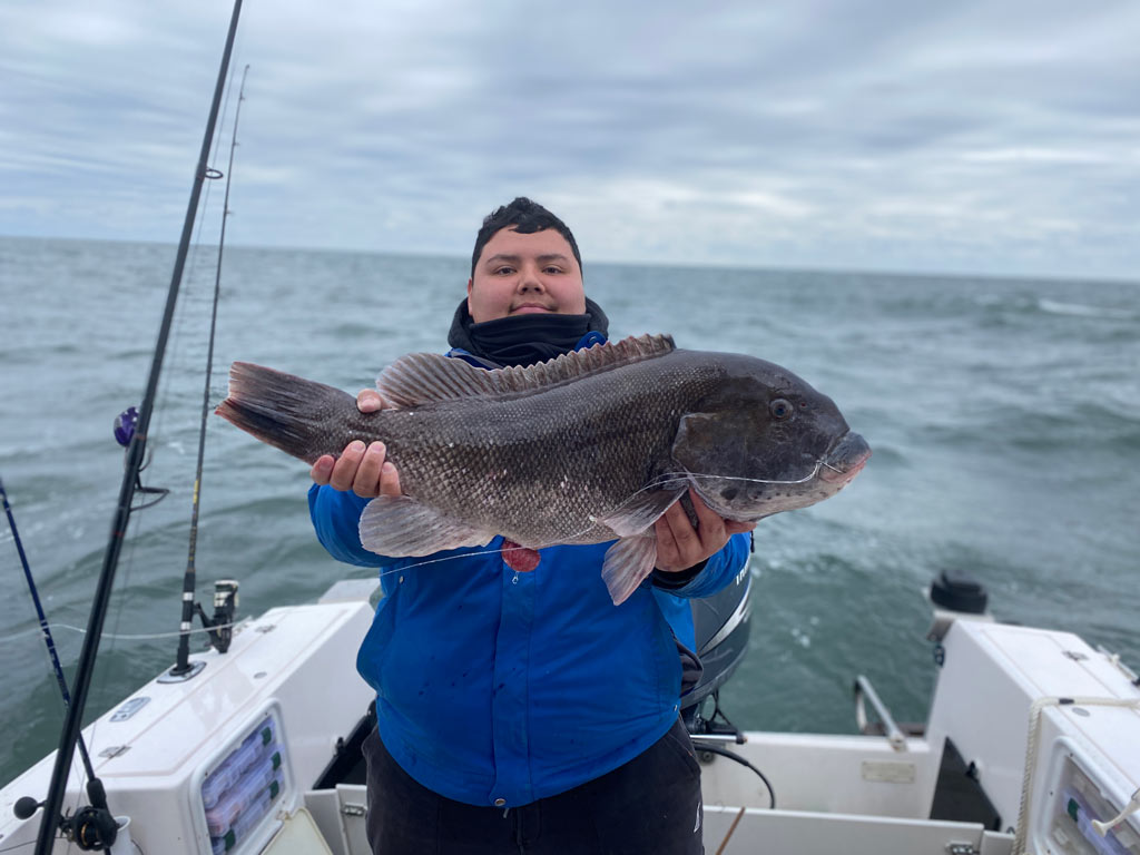 A boy standing on a boat, holding a sizeable Tautog he caught.