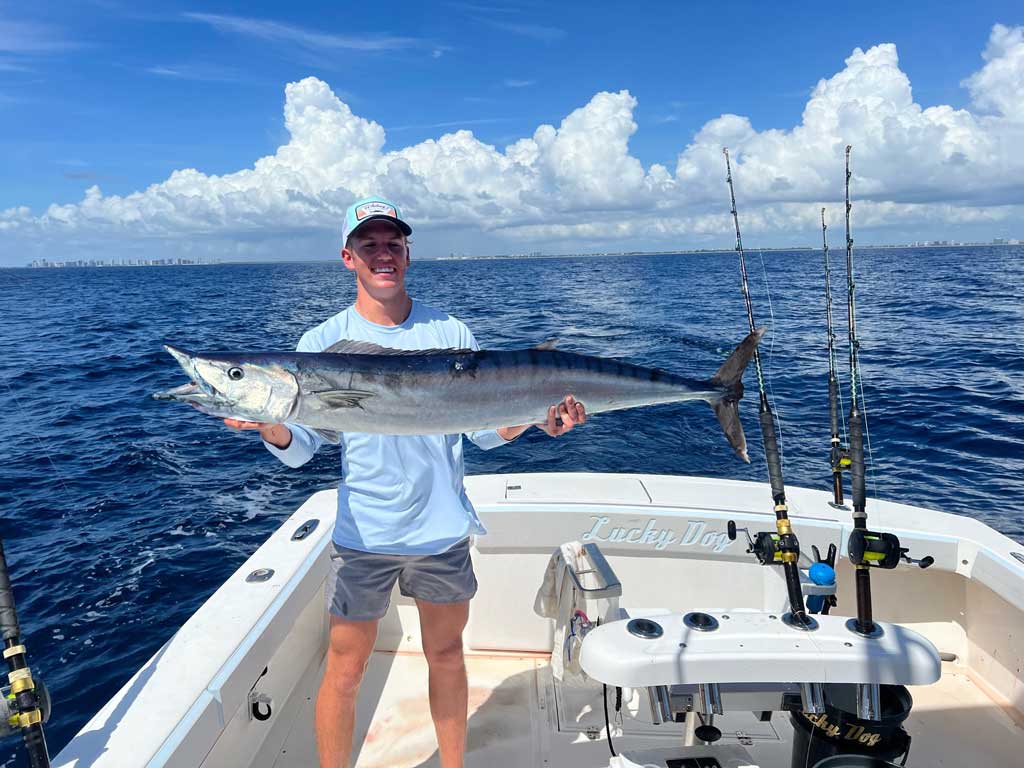 A man standing on a boat, posing with a big Wahoo he caught.