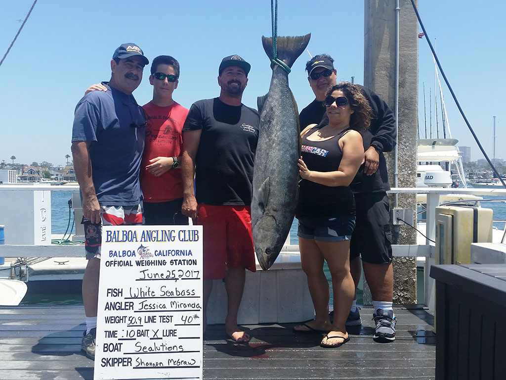 A group of anglers pose on the dock with a large White Seabass that's hanging next to a board indicating its weight and size