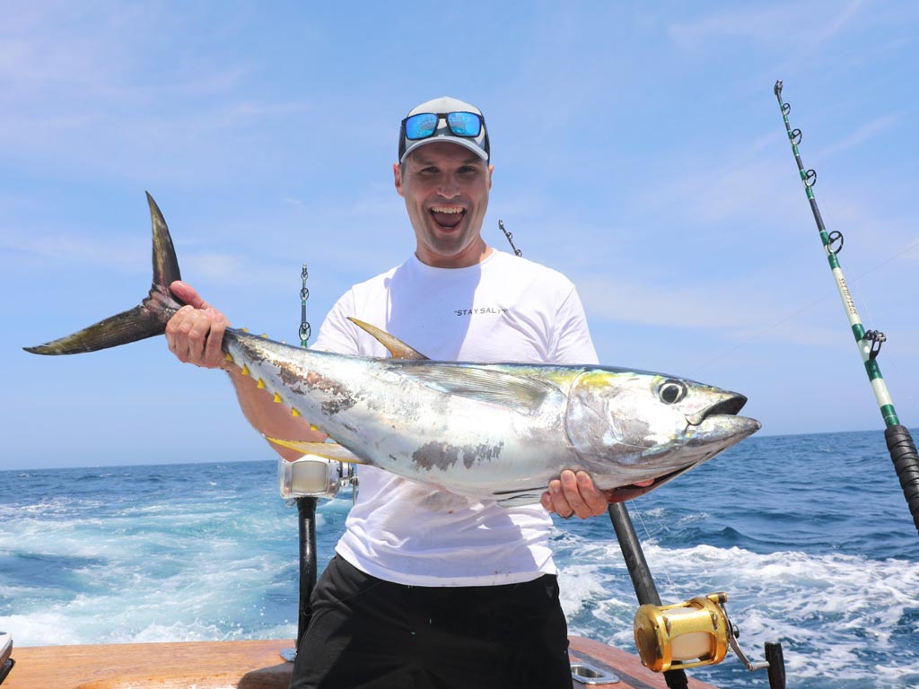 A smiling angler posing with a Yellowfin Tuna he reeled in, with a few fishing rods and the ocean behind him.