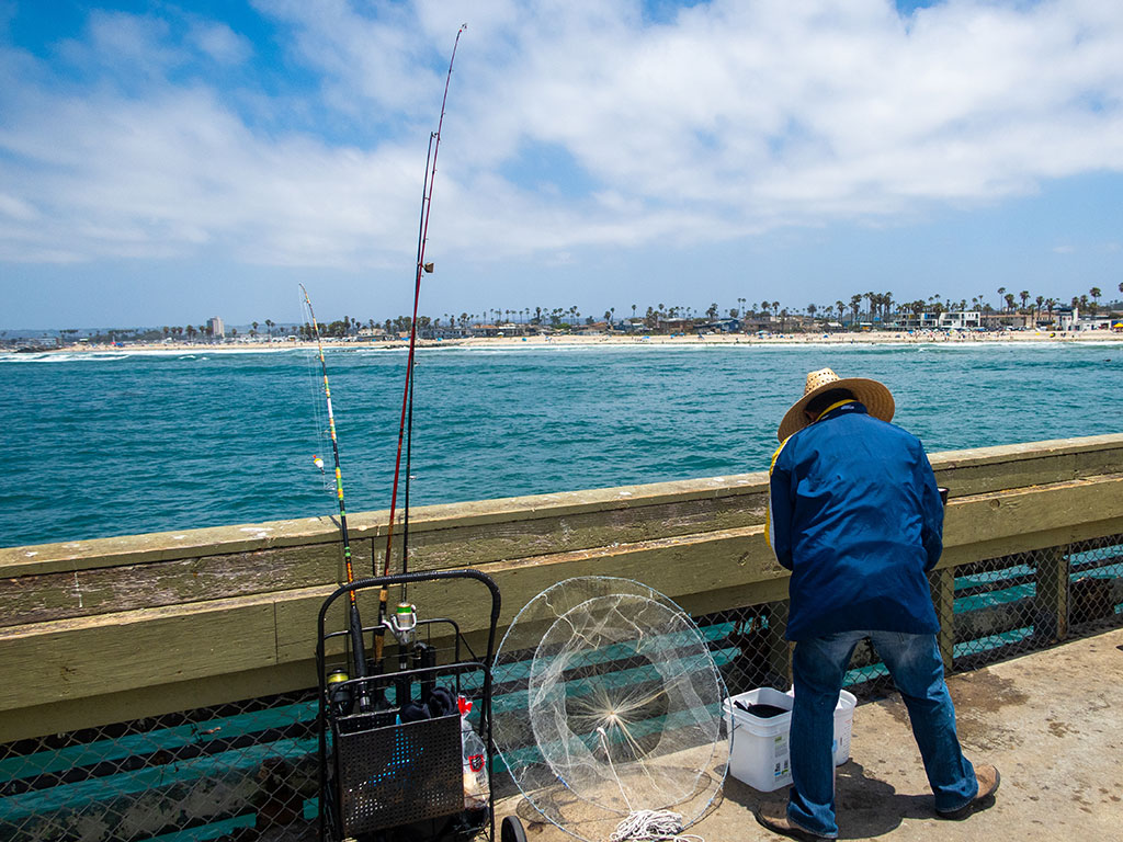 A rearview shot of an angler bending down on a fishing pier with a rod to his left, with land in the background