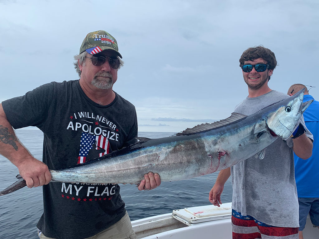 Two fishermen standing on a fishing boat, holding a Wahoo on a cloudy day