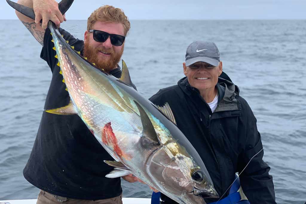 Two smiling anglers on a boat, one holding a big Yellowfin Tuna, the other standing next to him