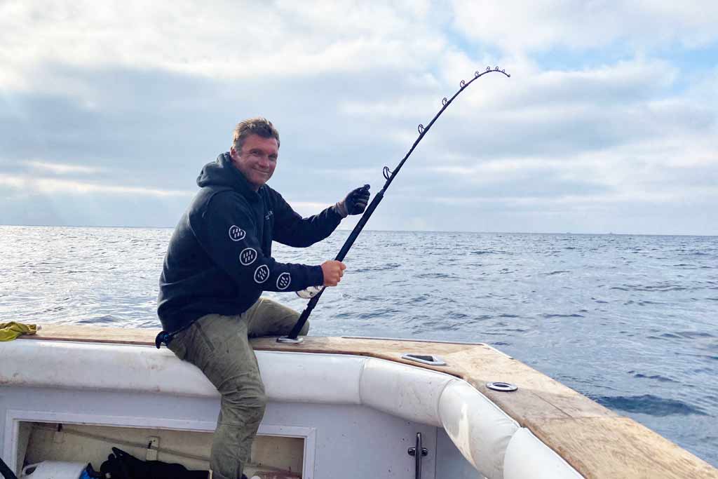 A fisherman sitting on a boat rail with one leg in and one leg out of the boat, holding a bent rod in front of him