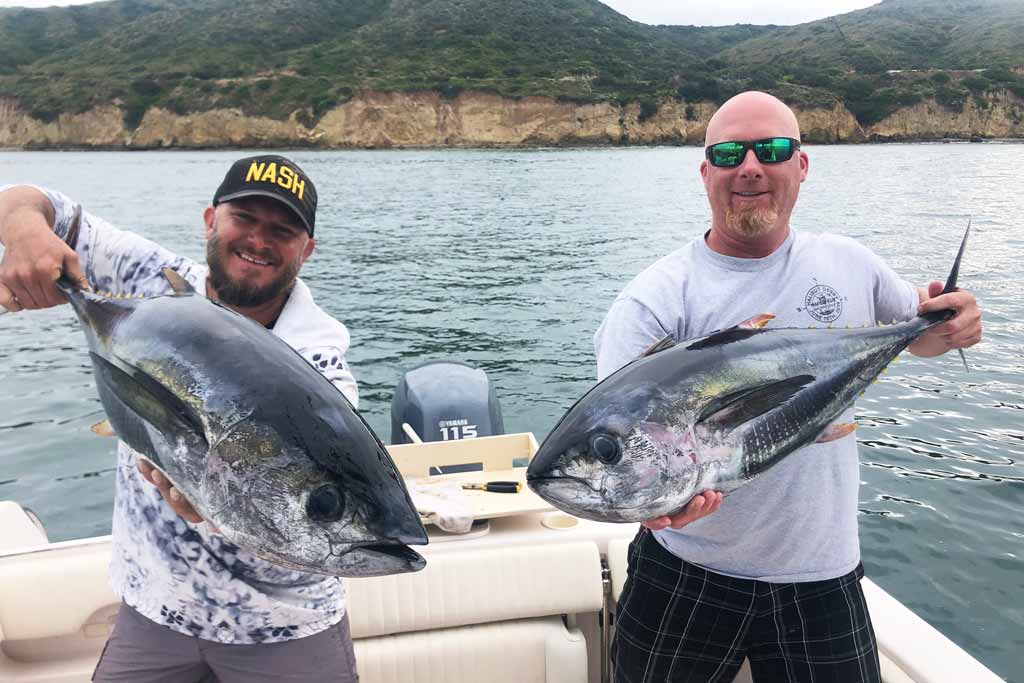 Two smiling anglers on a fishing boat, each holding a Bluefin Tuna with land behind them