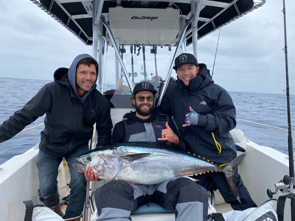 An angler sitting on a boat with a Bluefin Tuna in his lap, with two of his friends standing on either side of him.