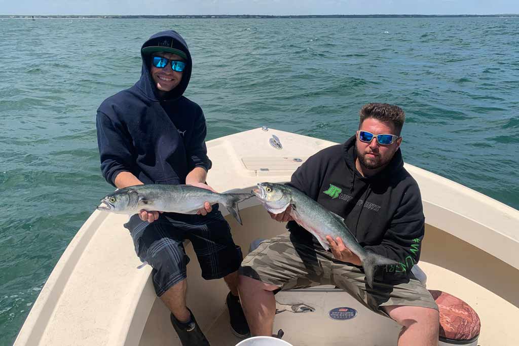 Two men sitting on a boat, holding a Bluefish each with the water behind them