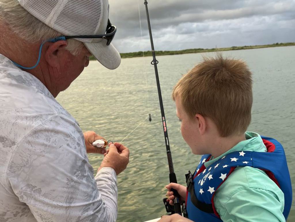A captain demonstrating how to prepare bait and as a kid carefully watches while holding a fishing rod and getting ready to try fishing