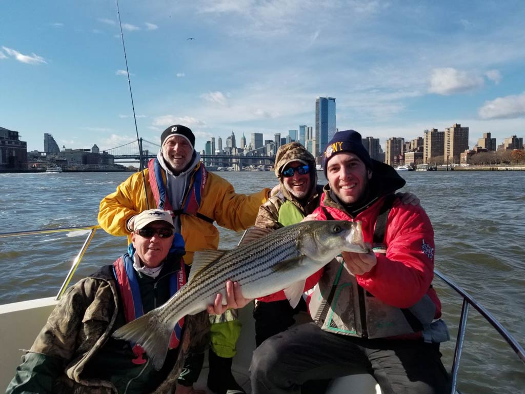 A group photo of friends posing against the skyline of the city with Striped Bass they caught while fishing in New York City during fall