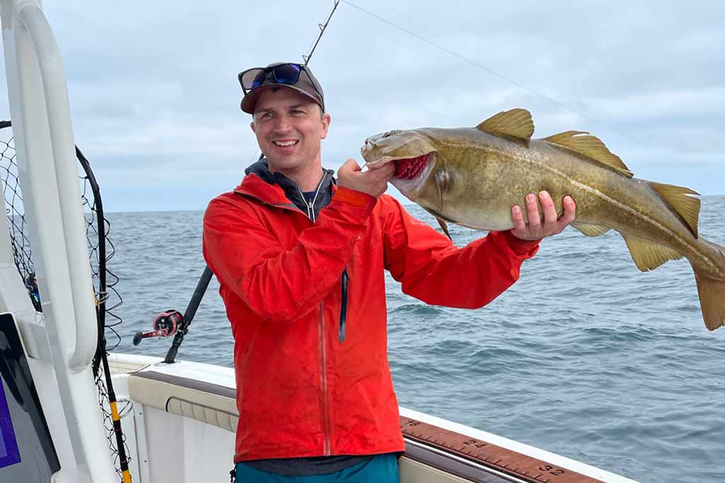 A smiling man standing on a boat, holding a big Cod fish, with water and cloudy skies in the background