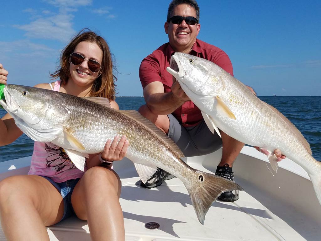 A photo of two anglers on a charter fishing boat looking happy while holding their big Redfish catches with both hands