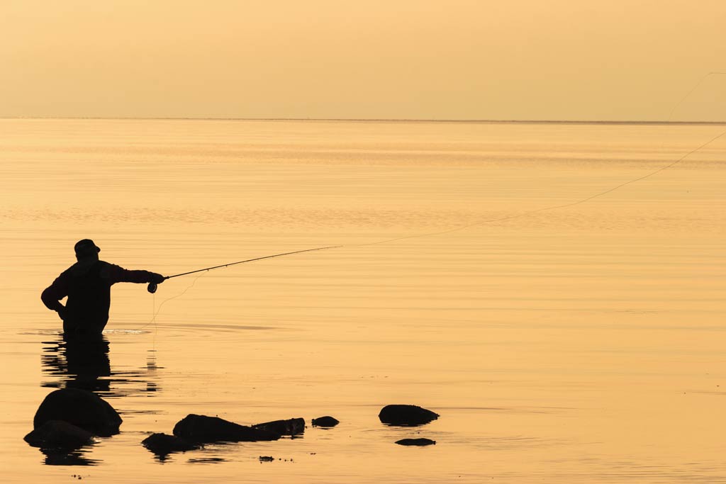 A fly fisherman wading in the surf, holding a fly rod, at sunset