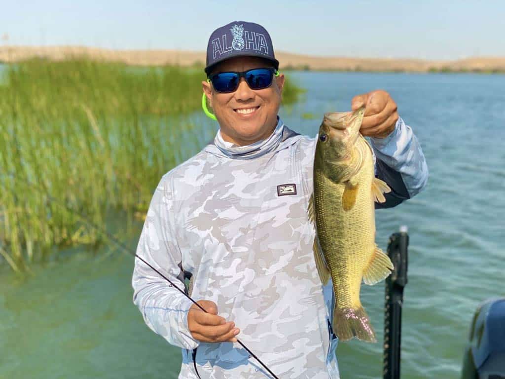 An excellent photo of an angler holding Bass in one hand and fly fishing gear in the other while standing on a charter fishing boat in the California Delta