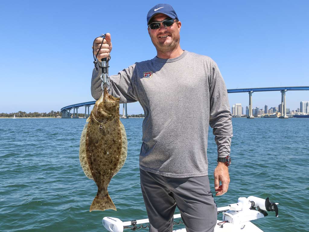 An angler standing on a boat, posing with a decent-sized Halibut he caught while fishing off of Point Loma.