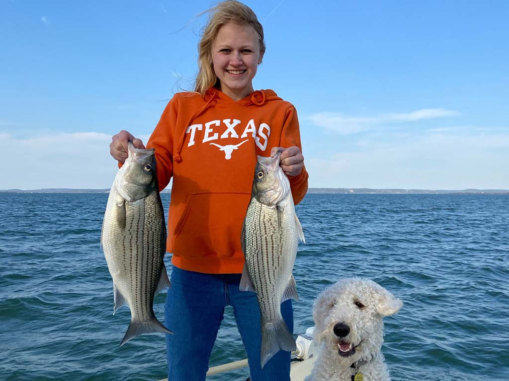 A woman standing on a boat, holding two Hybrid Bass she caught, with her dog next to her.