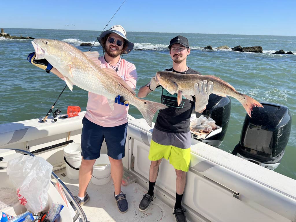 Two satisfied anglers standing on a charter fishing boat against a background of jetties and rocks, while showing off their Redfish catches