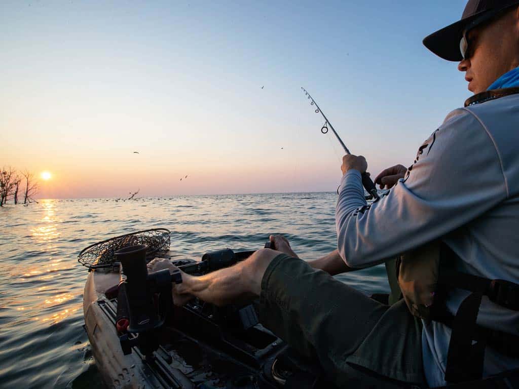 A sunset view of an angler fishing from his kayak with trees sticking out of the water at sunset