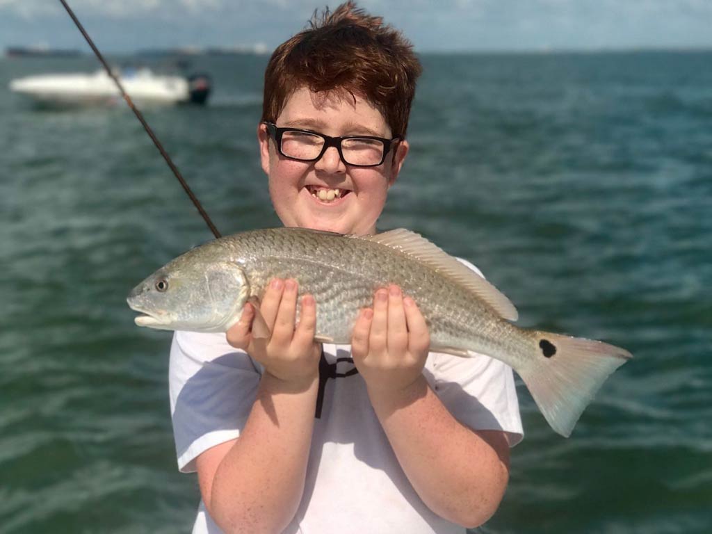 A cute photo of a kid smiling and holding small Redfish with both his hands while standing on a boat