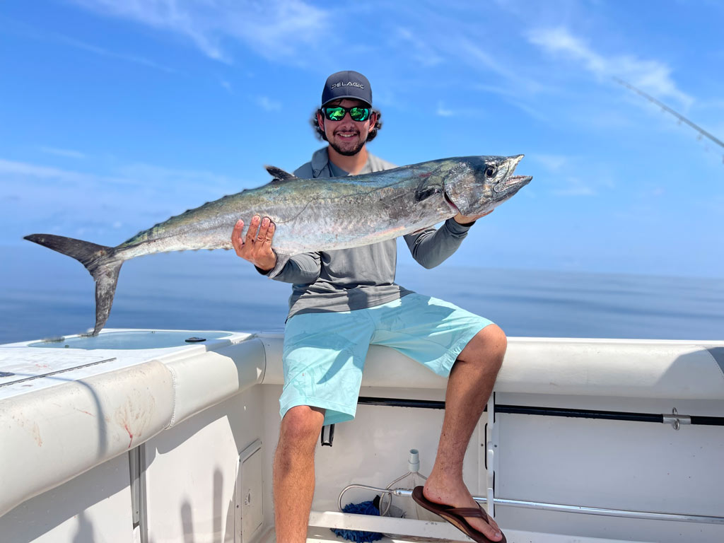 An angler sitting on side of the boat, holding a huge King Mackerel he reeled in.