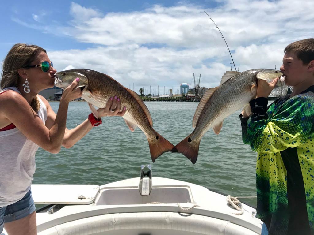 A woman and a teenage boy kissing a Redfish each that they caught while standing on a charter boat after the trip