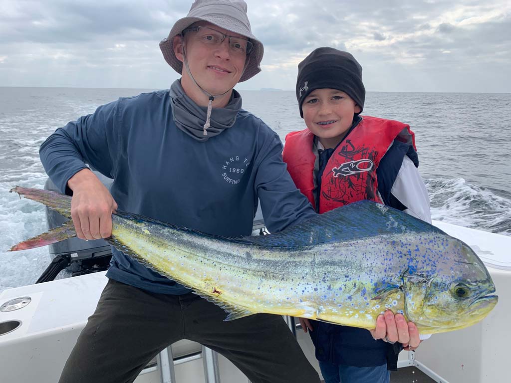 A boy and a man sitting on the back deck of a boat, holding a big Mahi Mahi caught fishing offshore from Point Loma.