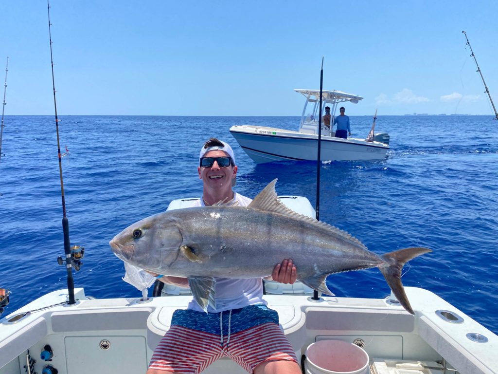 A smiling angler holding a large Amberjack while sitting on a charter fishing boat with another boat behind him in Miami Beach, Florida