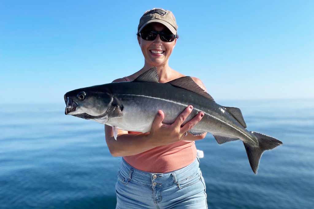 A smiling woman in a cap and sunglasses holding a Pollock fish, with blue skies and water in the background