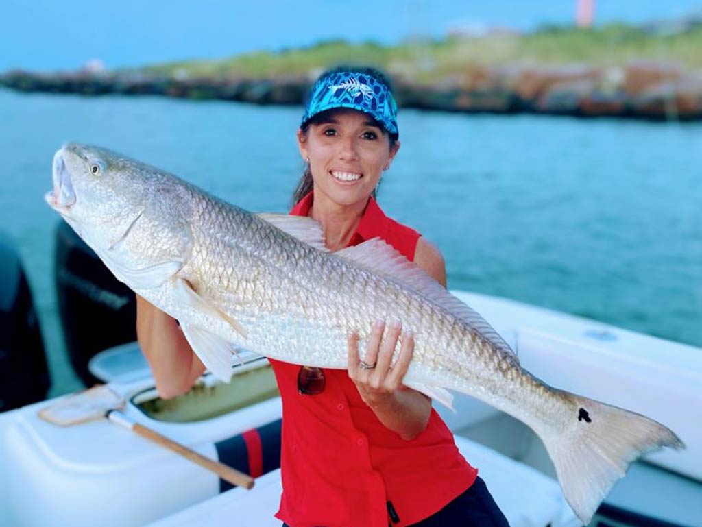 A woman posing against the shoreline in the background and holding a Redfish with both hands while standing on a Galveston fishing charter boat