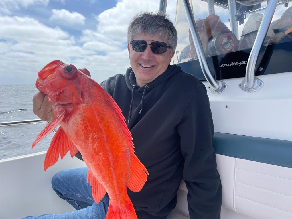 An angler squatting against the center console of a boat, holding a Rockfish in his right hand.