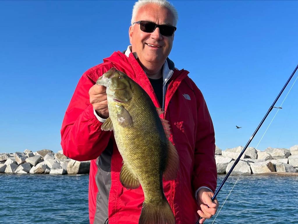 A photo of an angler in a red jacket standing against the rocky shoreline in the background and holding Bass caught while shore casting in New York City