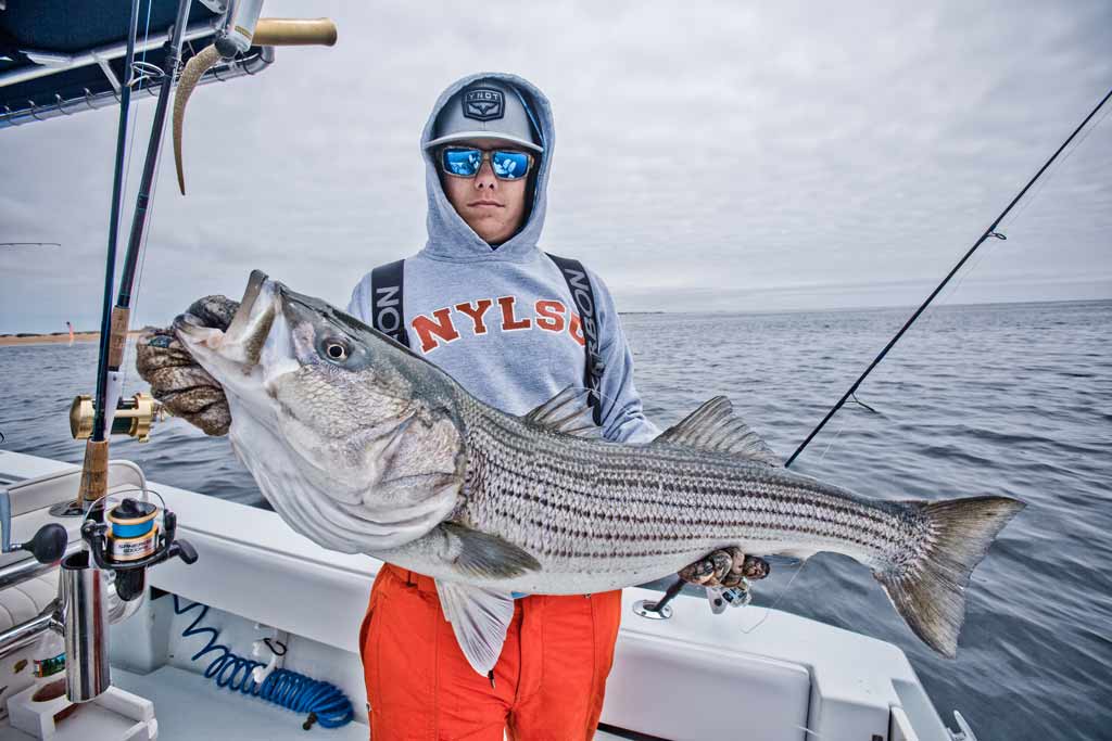 A young angler in a cap and sunglasses standing on a charter boat, holding a big Striped Bass